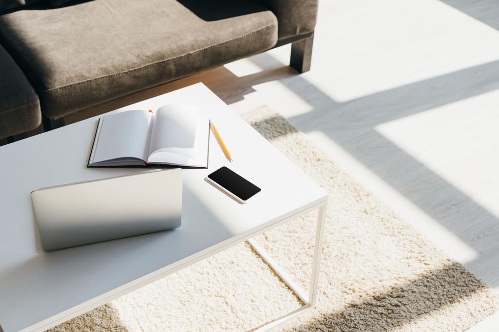 гостиная с диваном и столом с ноутбуком, смартфоном, блокнотом и карандашом в солнечном свете
 - Фото, изображение