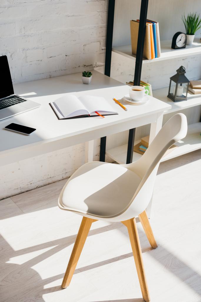 γραφείο στο σπίτι με smartphone, φλιτζάνι καφέ και φορητό υπολογιστή στο τραπέζι στο φως του ήλιου - Φωτογραφία, εικόνα