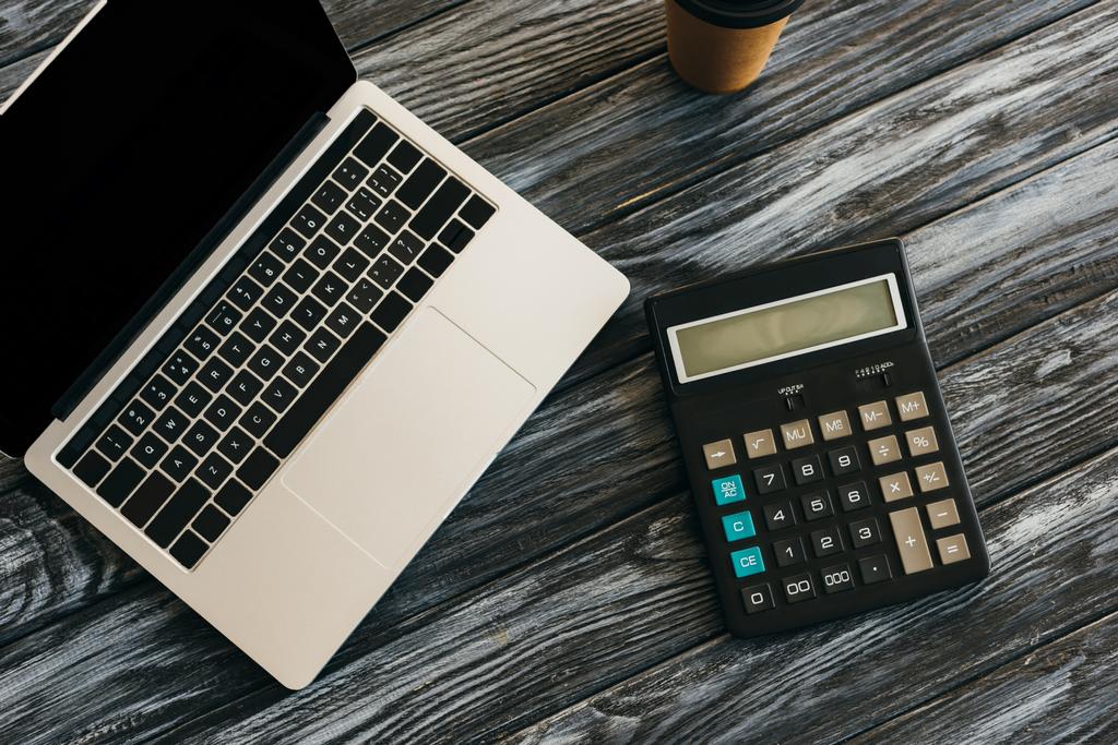 vrchní pohled na kalkulačku, kávu a notebook s prázdnou obrazovkou na tmavém dřevěném povrchu - Fotografie, Obrázek