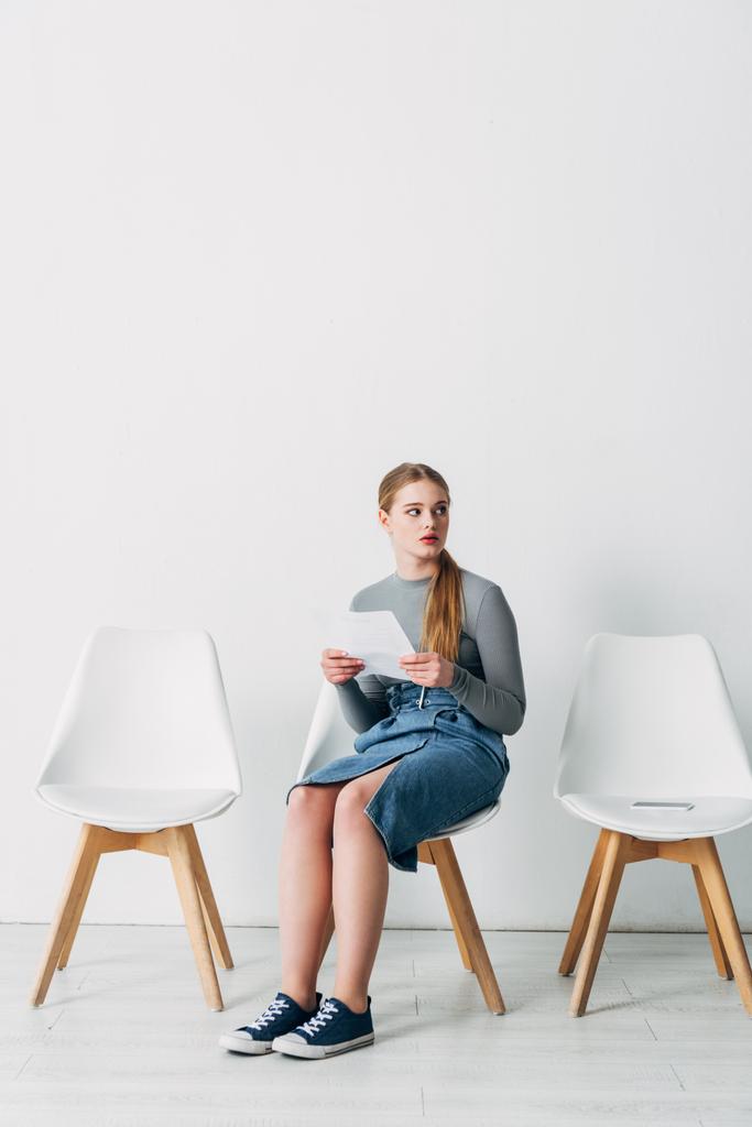Όμορφη υπάλληλος με βιογραφικό κοιτάζοντας μακριά κοντά smartphone σε καρέκλα στο γραφείο  - Φωτογραφία, εικόνα