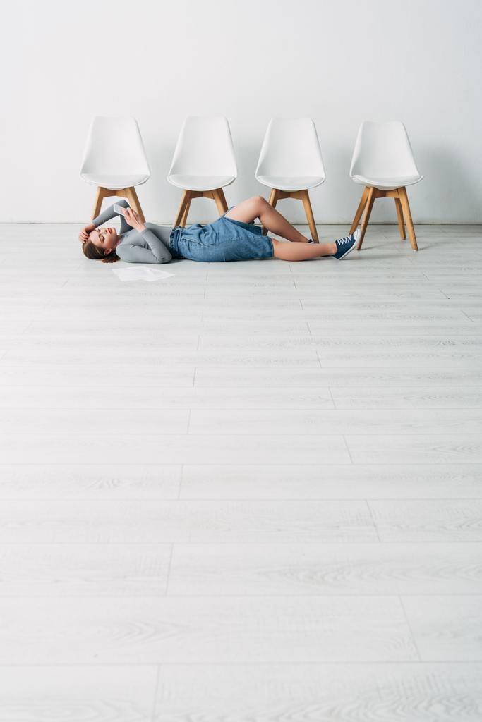Niedrigwinkel-Ansicht attraktiver Mitarbeiter mit Smartphone, während sie im Büro in der Nähe des Lebenslaufs auf dem Boden liegen  - Foto, Bild