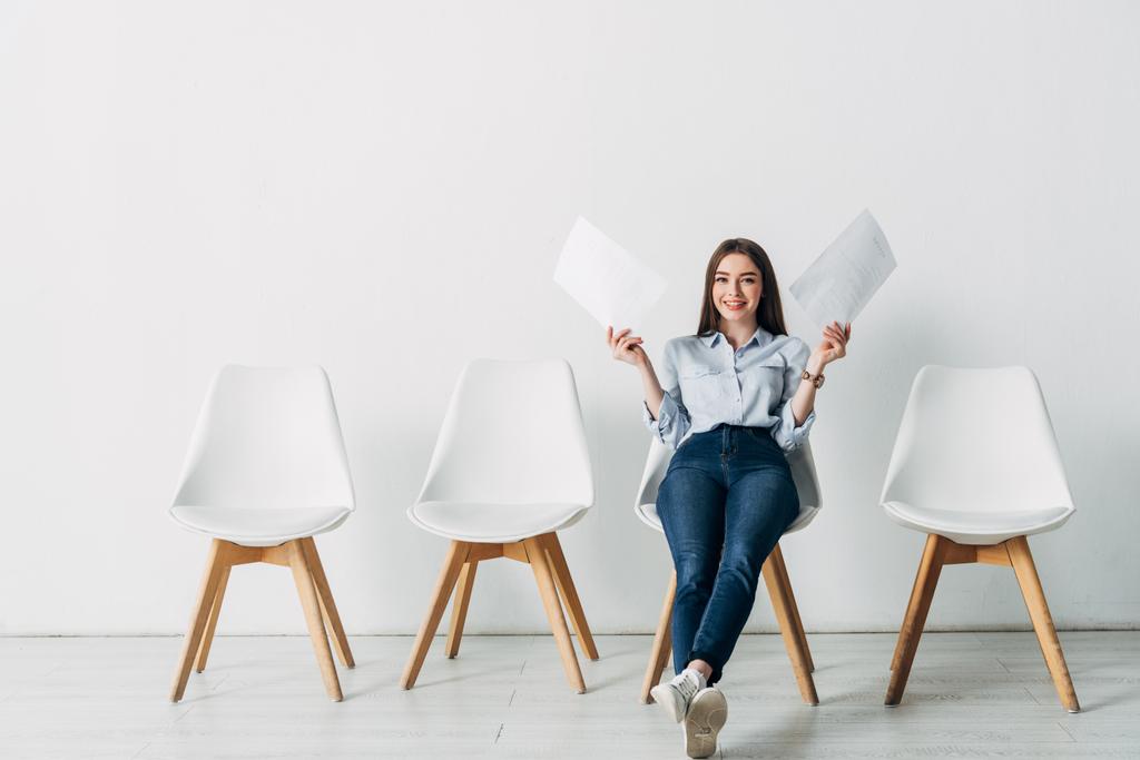 Smiling woman holding resume and smiling at camera on chair in office  - Photo, Image