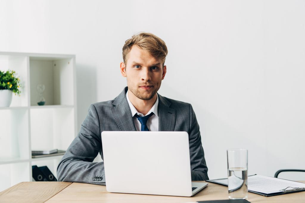 Handsome recruiter looking at camera near laptop and clipboard on table  - Photo, Image