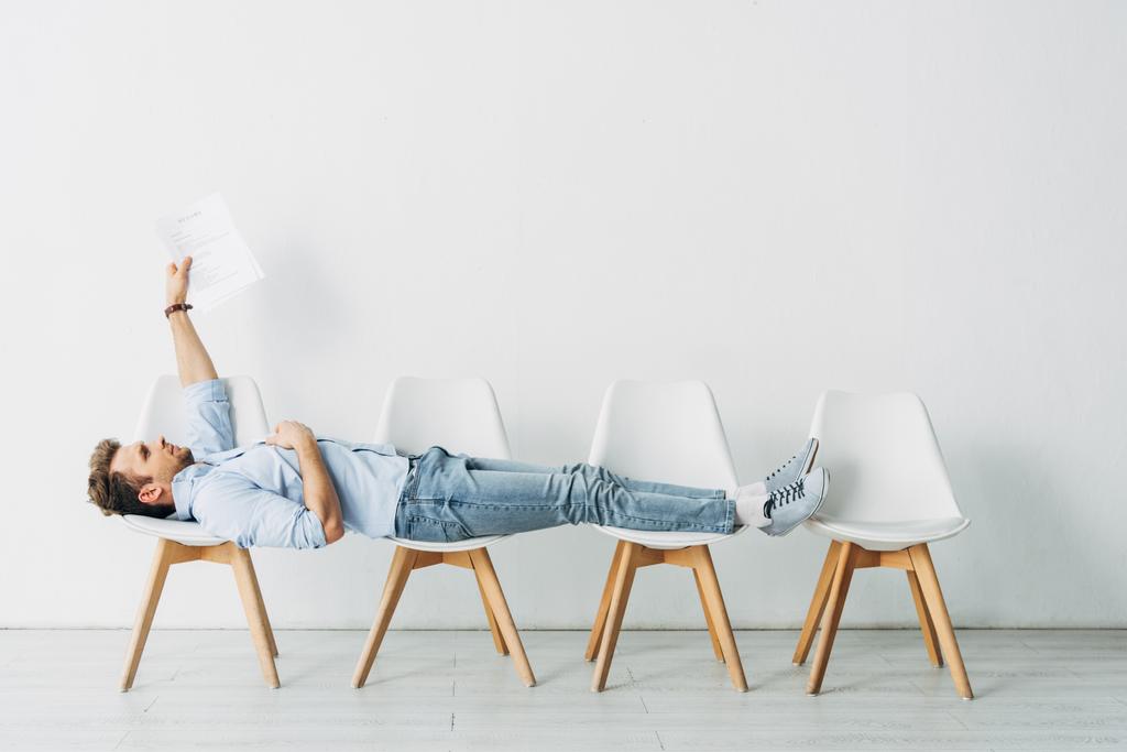 Side view of employee holding resume while lying on chairs in office  - Photo, Image