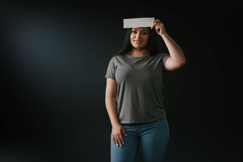 Front view of plus size plus beautiful girl with epmty card on forehead on black background - Photo, Image