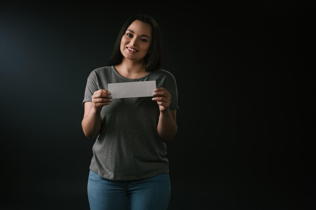 Front view of positive plus size girl holding empty card on black background - Photo, Image