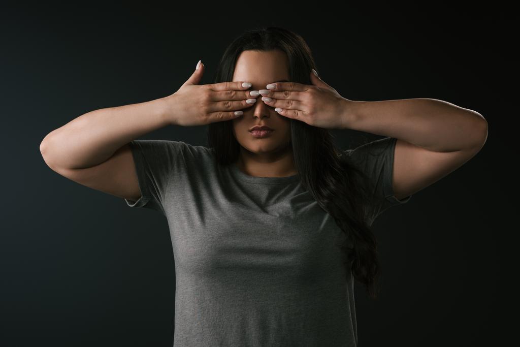 Vorderseite des Plus-Size-Mädchens, das Blindheit ausübt, indem es die Augen mit den Händen auf schwarzem Hintergrund verdeckt  - Foto, Bild