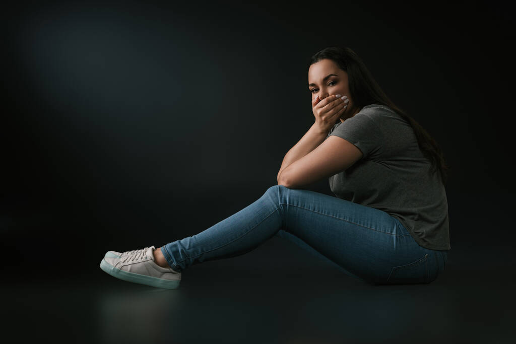 Side view of beautiful plus size girl covering mouth with hand and sitting on black background - Photo, Image