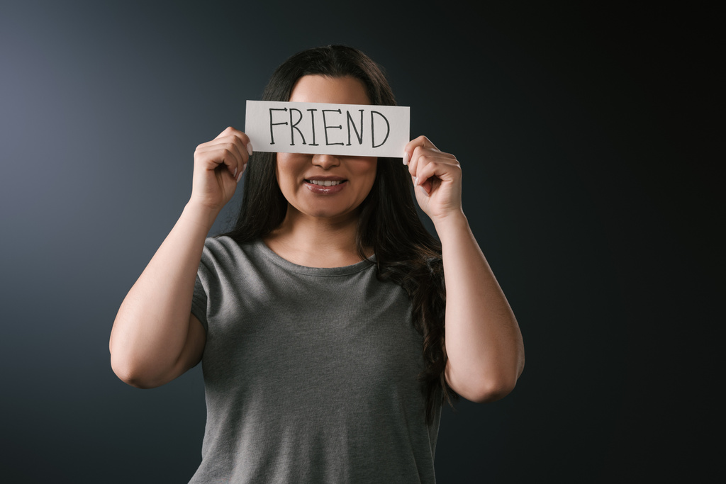 Vista frontal de la sonriente chica de tamaño grande cubriendo la cara con tarjeta con la palabra amigo sobre fondo negro
 - Foto, Imagen