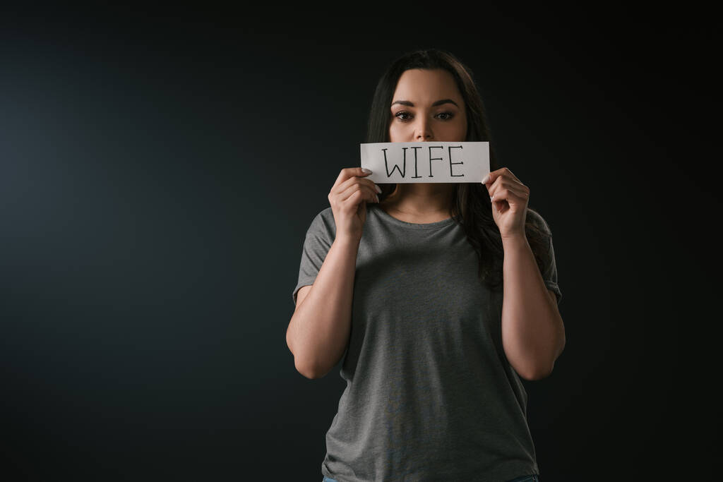 Front view of serious plus size girl holding card with word wife on black background - Photo, Image