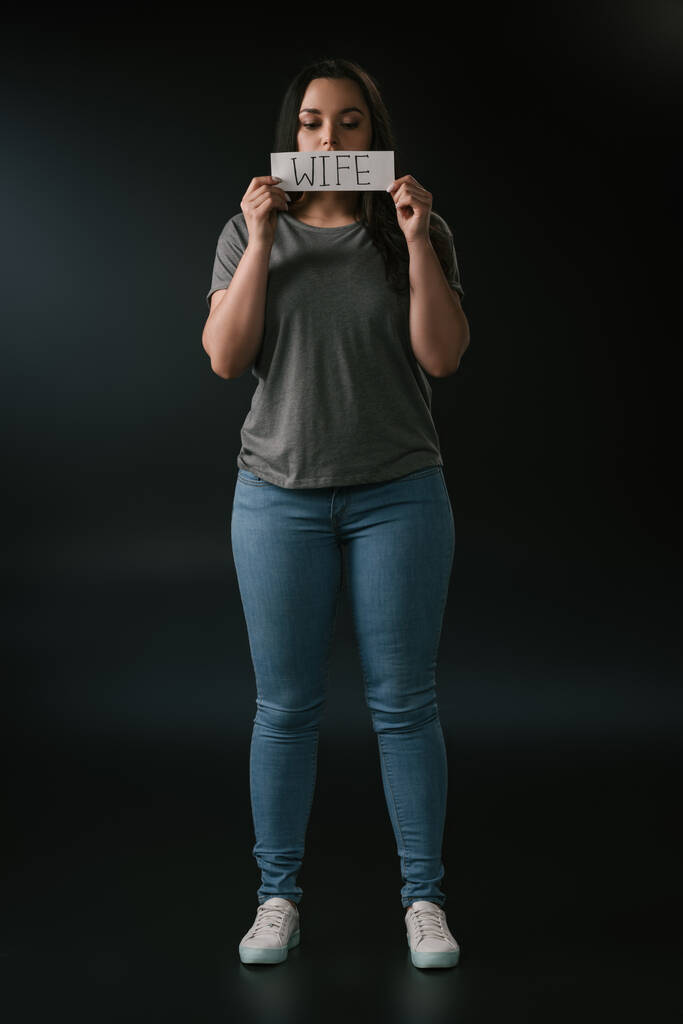 Полный вид девушки plus size, держащей открытку со словом "жена" на черном фоне
 - Фото, изображение