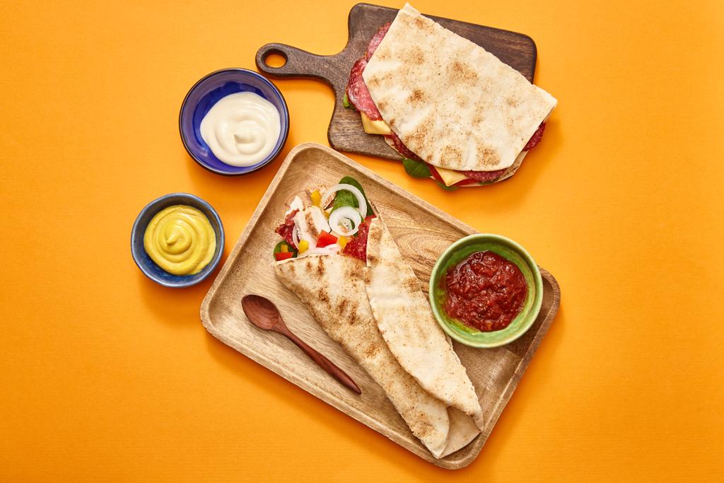 vrchní pohled na čerstvý sendvič se salámem v pita a burrito s kuřecím masem na deskách v blízkosti omáčky na oranžovém pozadí - Fotografie, Obrázek