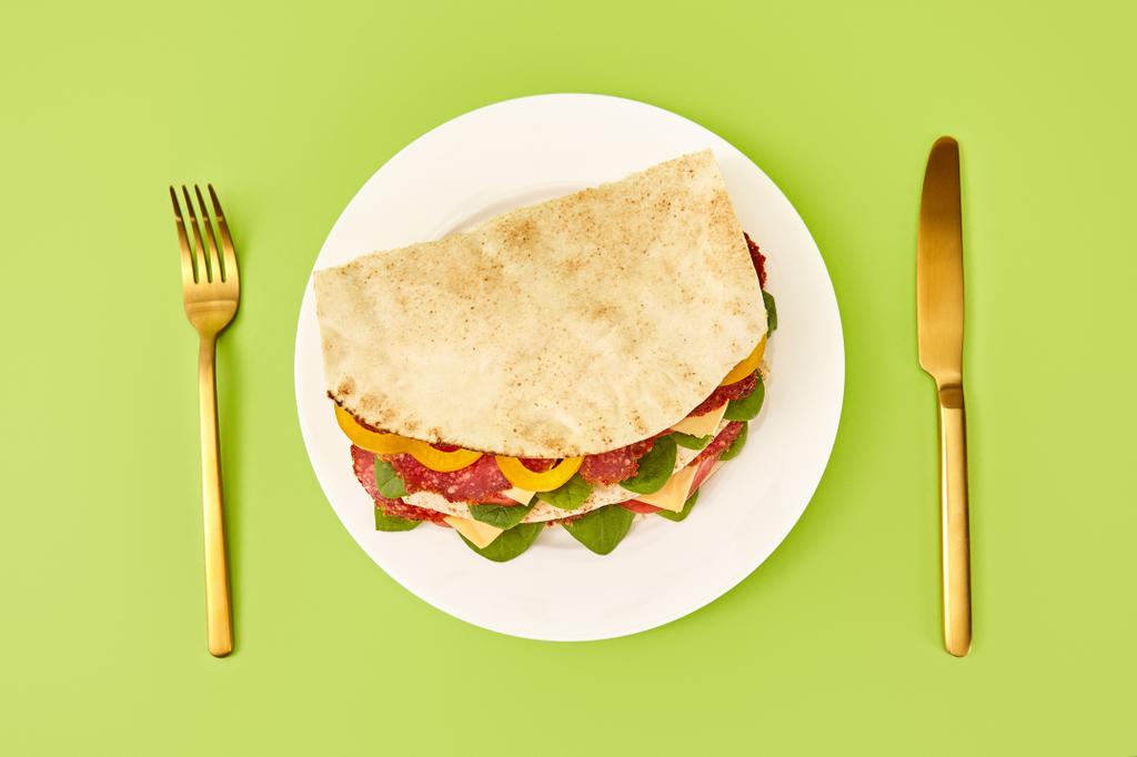 vista superior de sándwich fresco con salami, pita, verduras y queso servido en el plato cerca de tenedor de oro y cuchillo sobre fondo verde
 - Foto, imagen