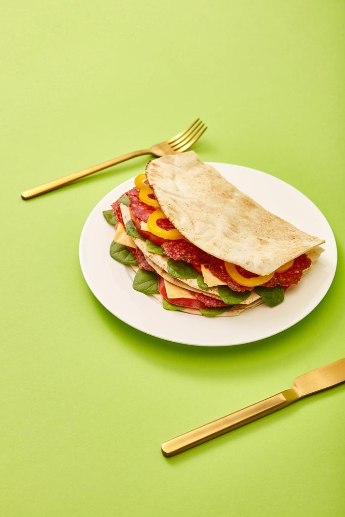 sándwich fresco con salami, pita, verduras y queso servido en el plato cerca de tenedor de oro y cuchillo sobre fondo verde
 - Foto, Imagen