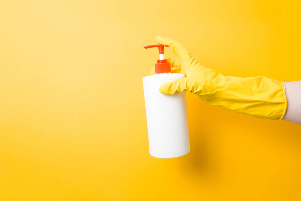 female hands in yellow rubber gloves holding a white soap dispenser and dishwashing and house cleaning products, yellow background, copy space, spring cleaning concept - Photo, Image