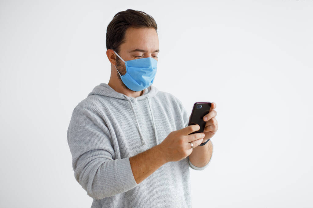 Bílý vousatý dospělý muž pomocí smartphonu, zatímco na sobě chirurgické masky izolované na bílém pozadí. Zdraví, epidemie, sociální média, komunikace a životní styl s kopírovacím prostorem - Fotografie, Obrázek
