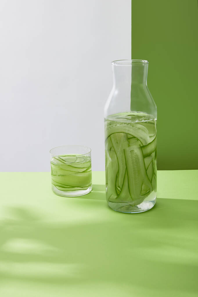 Bottle and glass with fresh drink made of sliced cucumbers on grey and green background - Photo, Image