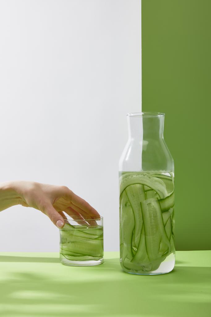 Dilimlenmiş salatalıktan yapılmış taze içecekle kadın eli değen cam kırıkları ve gri ve yeşil arka planda şişe. - Fotoğraf, Görsel