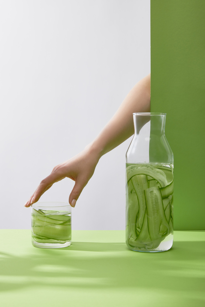 Gebonden zicht op vrouwelijk handaanraakglas met verse drank gemaakt van gesneden komkommers en fles op grijze en groene achtergrond - Foto, afbeelding