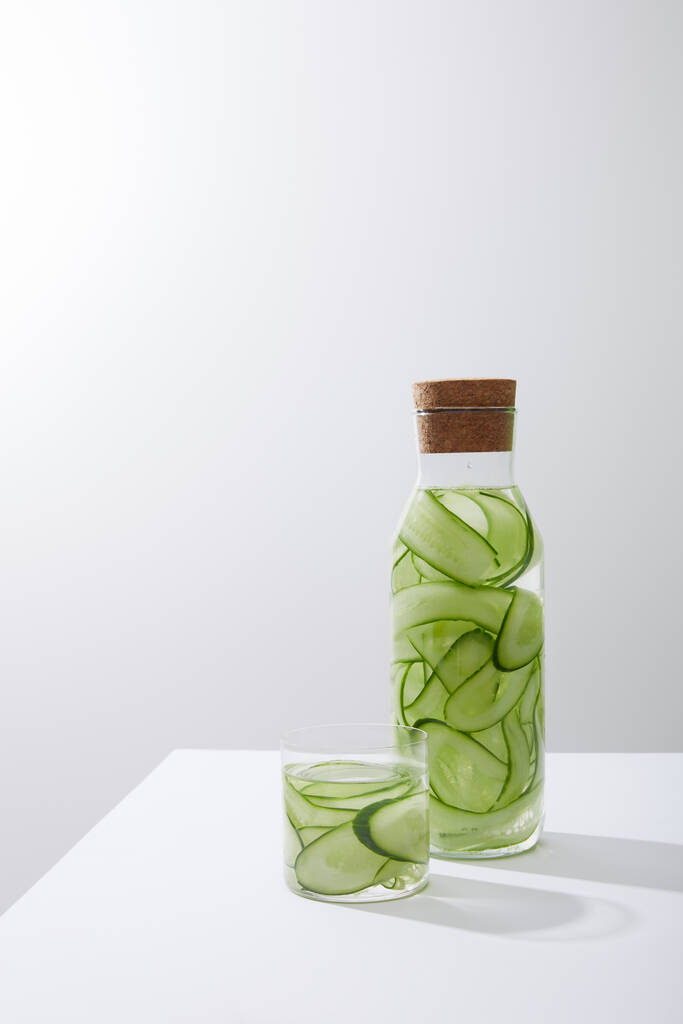 Стекло и бутылка с пробкой, наполненной водой и нарезанными огурцами на сером фоне
 - Фото, изображение