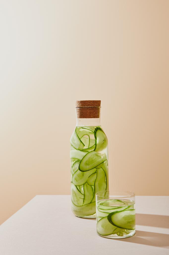 Glas en fles met kurk gevuld met water en gesneden komkommers op tafel op beige ondergrond - Foto, afbeelding