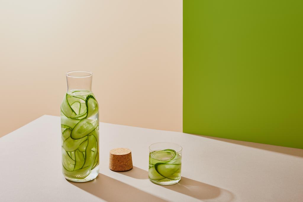 высокий угол обзора пробки, стекла и бутылки, наполненные водой и нарезанные огурцы на столе на бежевом и зеленом фоне
 - Фото, изображение
