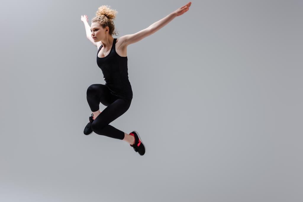 krullende sportvrouw met uitgestrekte handen springen geïsoleerd op grijs  - Foto, afbeelding