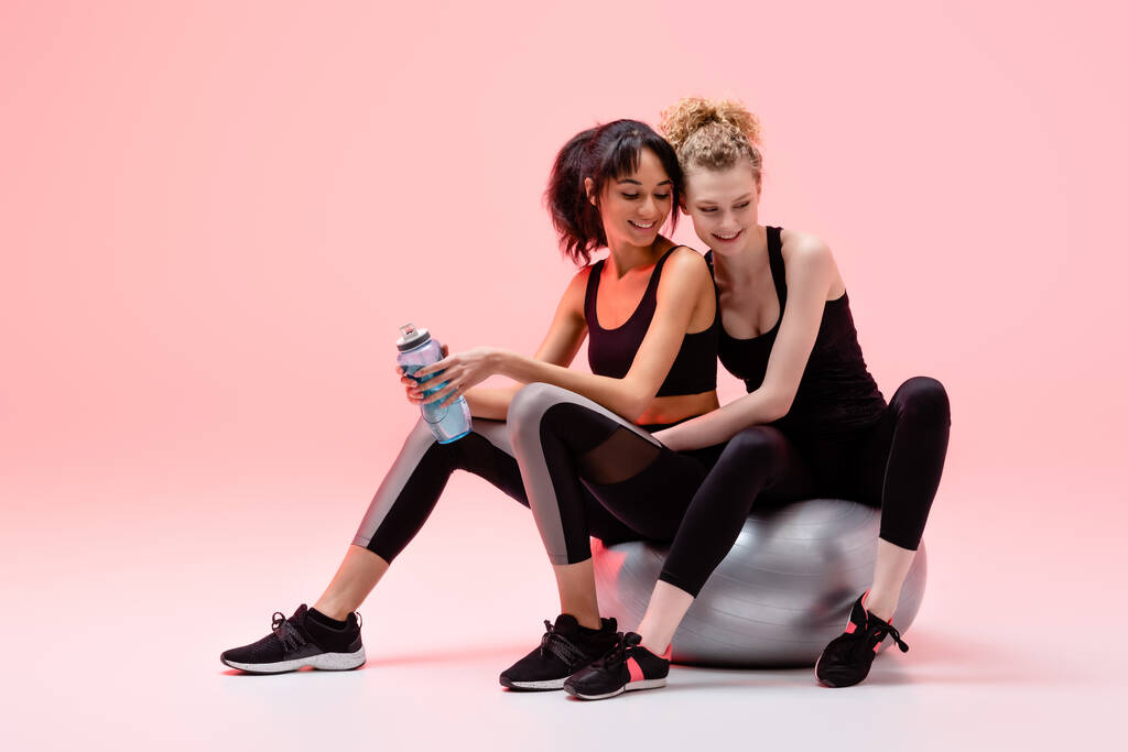 happy girl sitting on fitness mall with cheerful african american woman on pink  - Photo, Image