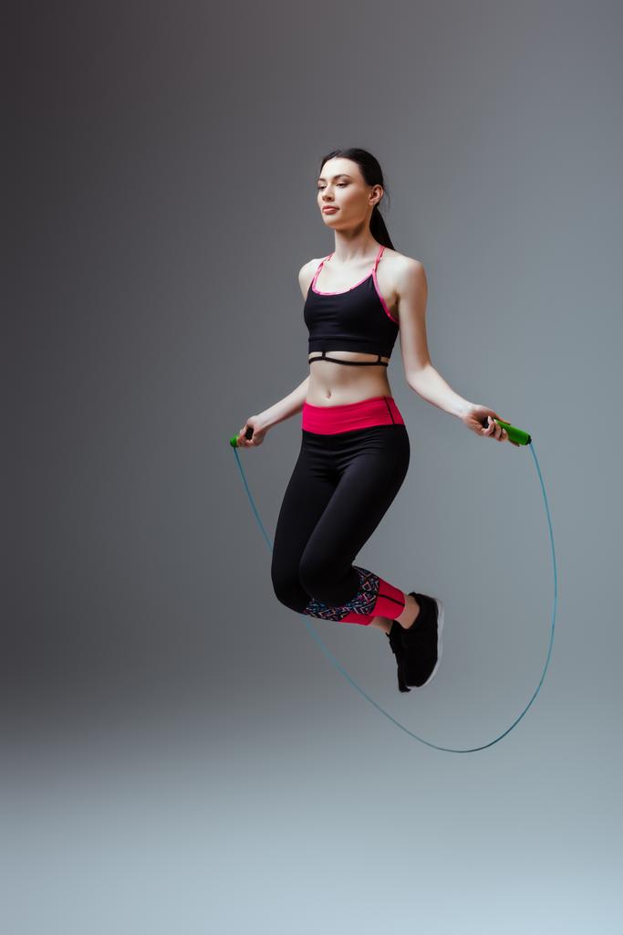 young woman in sportswear jumping with skipping rope on grey  - Photo, Image