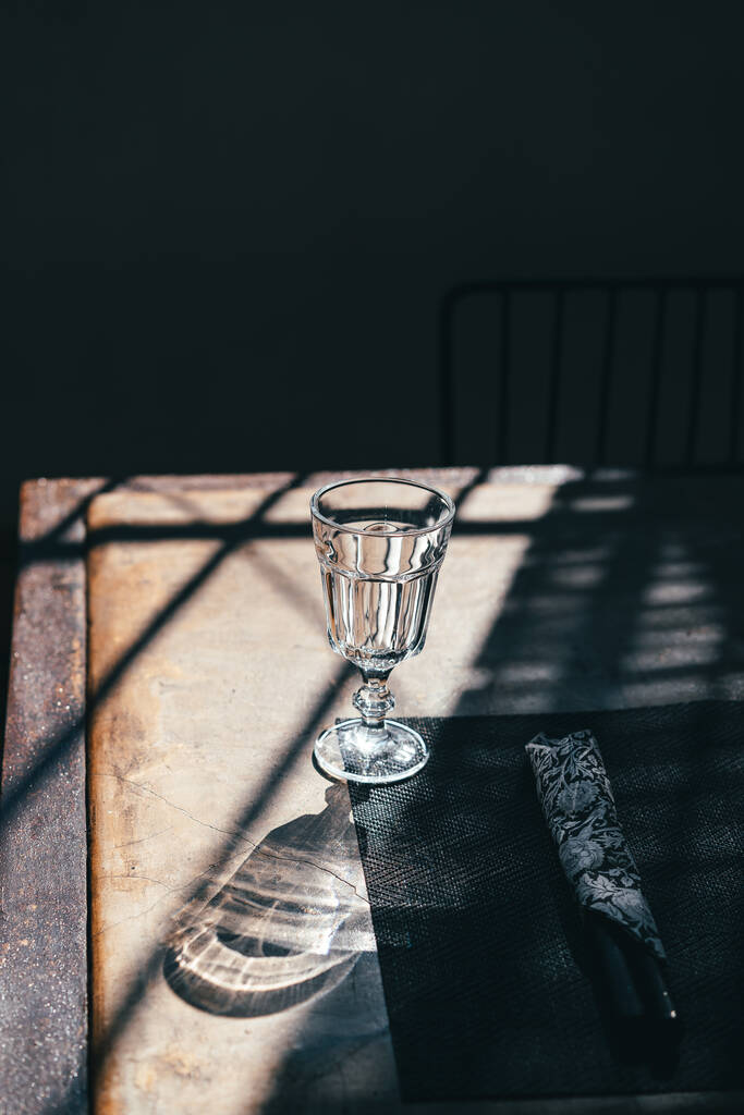 Ein gläserner Kelch auf einem Bein steht auf einem Tisch zwischen den Schatten im Sonnenlicht - Foto, Bild