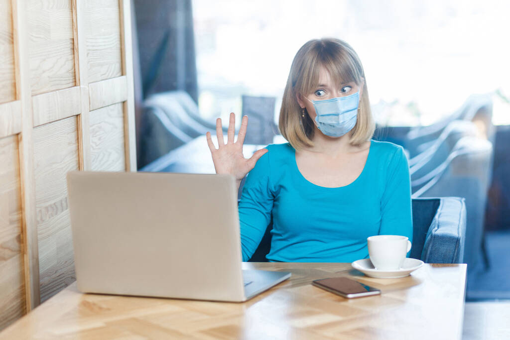Videoanruf, Online-Arbeit zu Hause. Porträt einer jungen Frau mit chirurgischer medizinischer Maske sitzt am Laptop und grüßt auf einer Videokonferenz. Raum-, Medizin- und Gesundheitskonzept - Foto, Bild