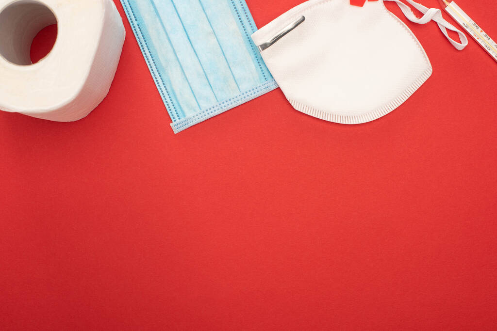 horní pohled na bílý toaletní papír, bezpečnostní a lékařské masky a teploměr na červeném pozadí - Fotografie, Obrázek