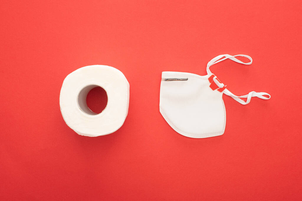 bovenaanzicht van wit schoon toiletpapier rol en veiligheidsmasker op rode achtergrond - Foto, afbeelding