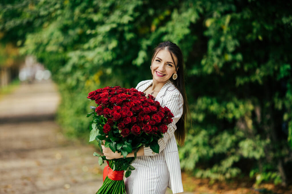 Модна розкішна сексуальна дівчина в елегантному костюмі, що тримає букет із сотень червоних троянд. Щасливий день Святого Валентина
 - Фото, зображення
