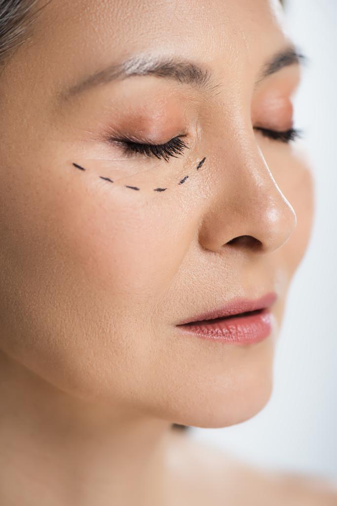 asiatische Frau mit geschlossenen Augen und plastischen Chirurgie Linien im Gesicht isoliert auf grau  - Foto, Bild