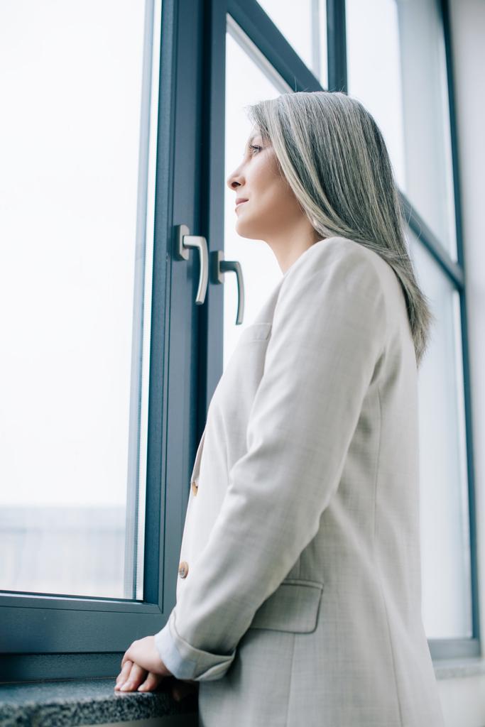 atractivo asiático mujer de negocios con gris pelo mirando a través de ventana
   - Foto, Imagen