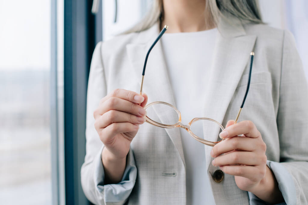 vista cortada de empresária executiva em terno cinza segurando óculos no escritório
 - Foto, Imagem