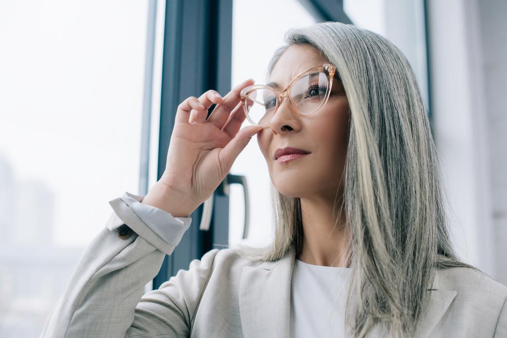 přemýšlivá asijská podnikatelka s šedými vlasy v šedém obleku a brýlemi v kanceláři - Fotografie, Obrázek