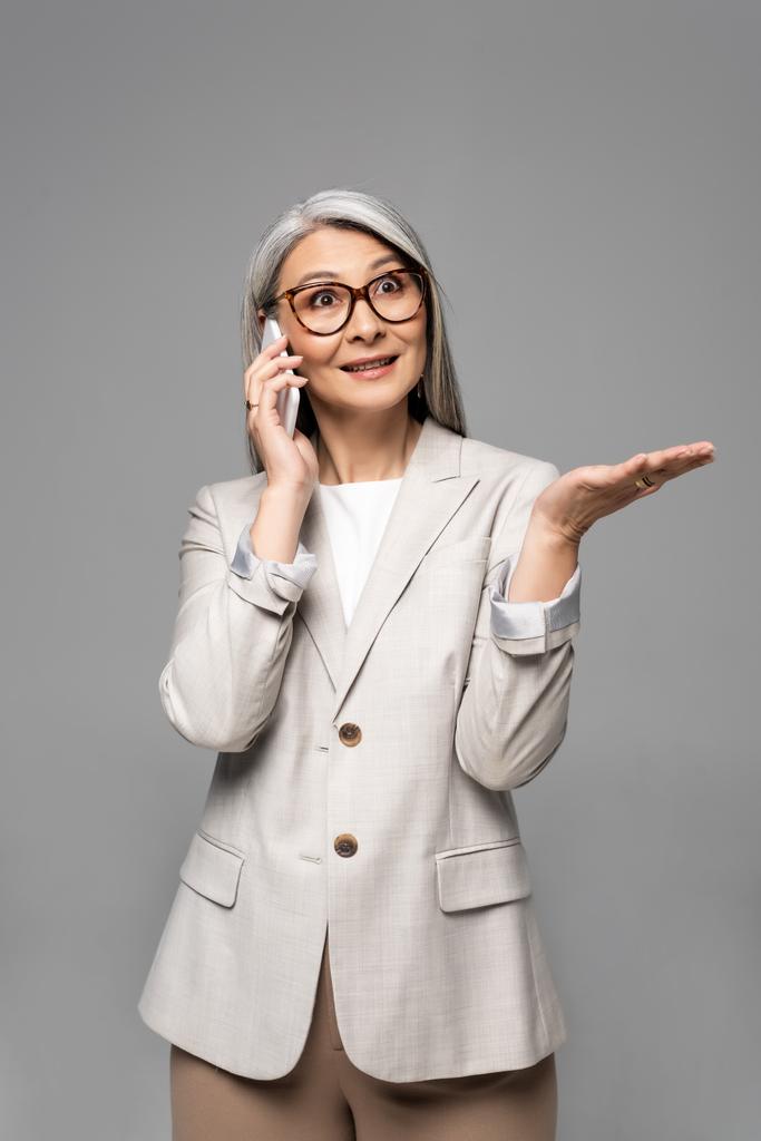 灰色の髪をした眼鏡のアジア系ビジネス女性が灰色で隔離されたスマートフォンで話しているのを見て - 写真・画像
