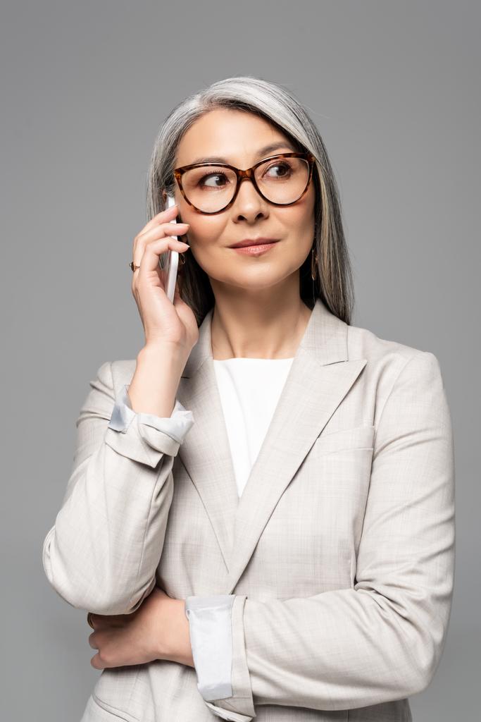 professional asian businesswoman in eyeglasses with grey hair talking on smartphone isolated on grey - Photo, Image