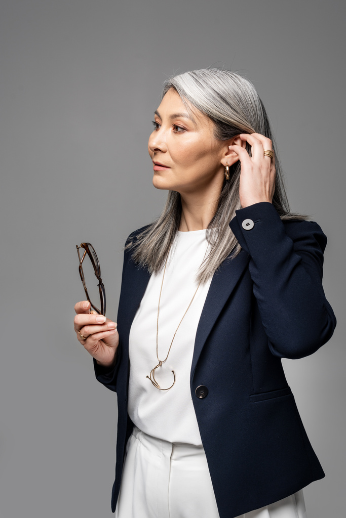 азіатська бізнес-леді з сірим волоссям тримає окуляри ізольовані на сірому
 - Фото, зображення