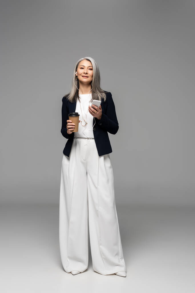 schöne asiatische Geschäftsfrau mit Kaffee zum Musikhören mit Kopfhörer und Smartphone auf grau - Foto, Bild