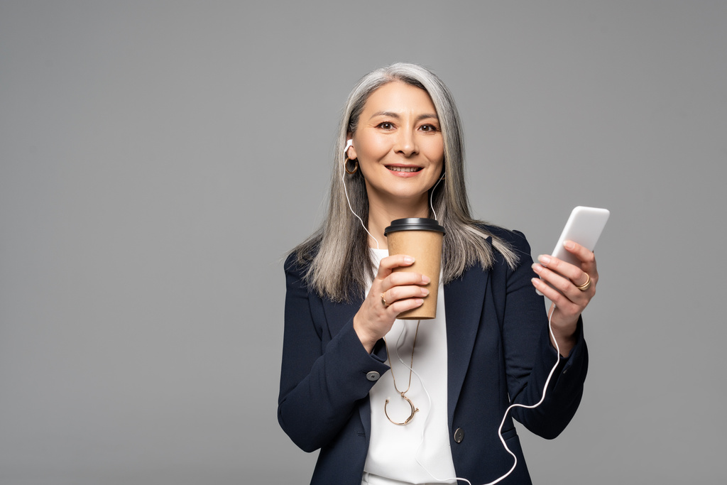 emotional asian businesswoman with coffee to go listening music with earphones and smartphone isolated on grey - Photo, Image