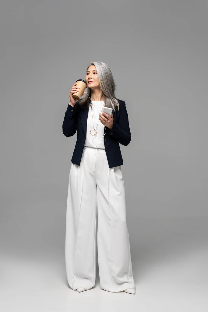 χαρούμενη Ασιάτισσα επιχειρηματίας με καφέ για να πάει να ακούσει μουσική με ακουστικά και smartphone σε γκρι - Φωτογραφία, εικόνα
