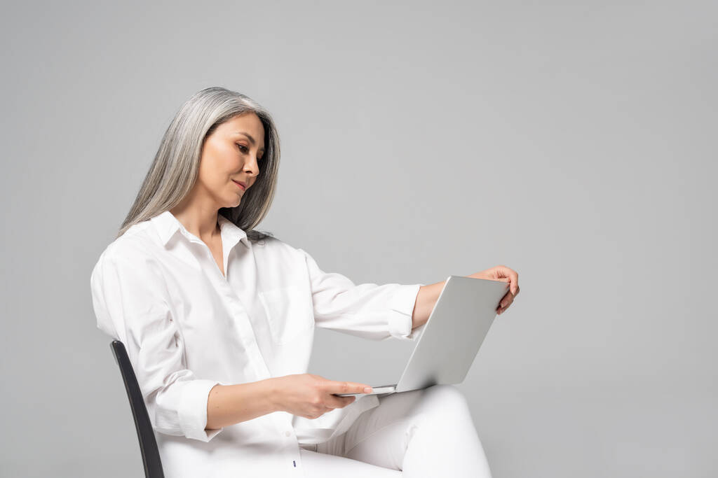 Erwachsene asiatische Frau mit grauen Haaren sitzt auf Stuhl und benutzt Laptop isoliert auf grau - Foto, Bild