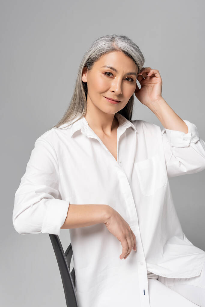 schöne nachdenkliche asiatische Frau mit grauen Haaren sitzt auf Stuhl isoliert auf grau - Foto, Bild