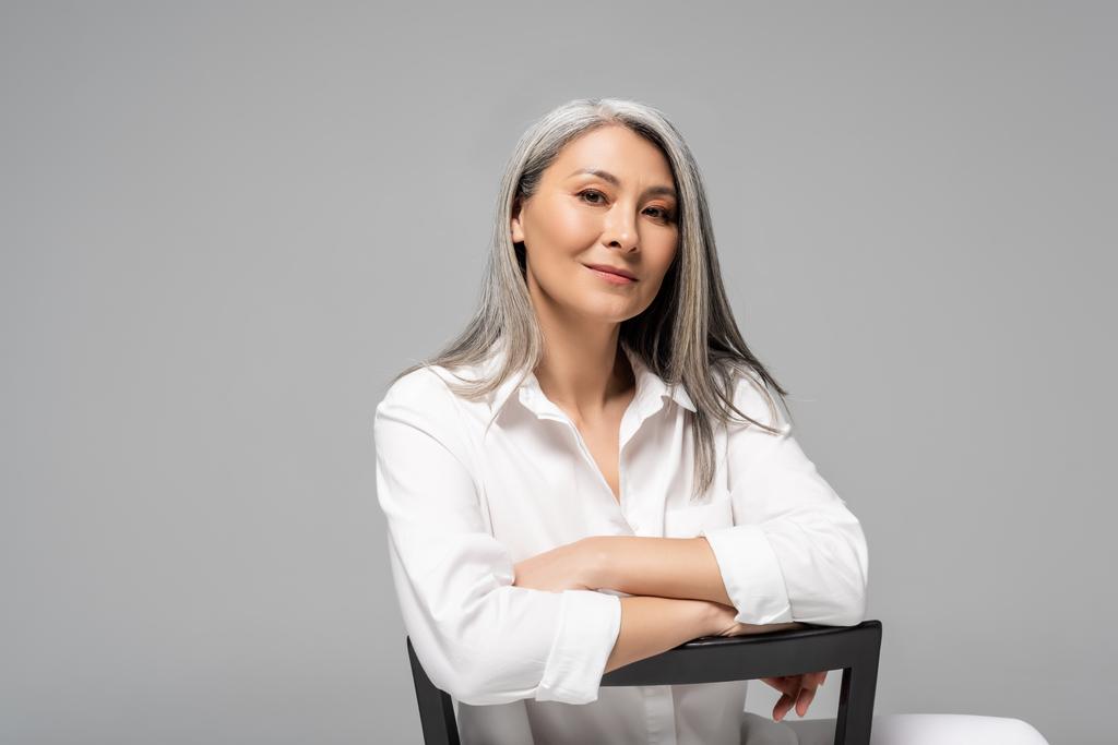 atrakcyjna Azji kobieta z siwymi włosami siedzi na krześle odizolowany na szary - Zdjęcie, obraz