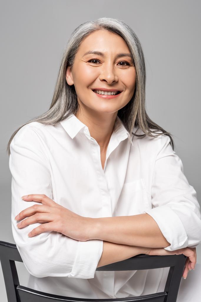 hermosa sonriente asiático mujer con gris pelo sentado en silla aislado en gris
 - Foto, imagen