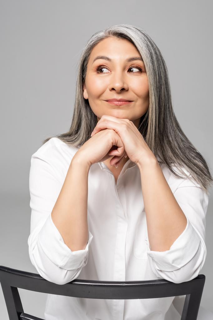 positive nachdenkliche asiatische Frau mit grauen Haaren sitzt auf Stuhl isoliert auf grau - Foto, Bild