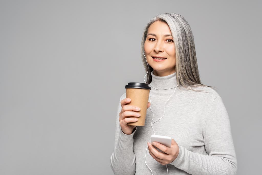 heureux asiatique femme dans col roulé avec café pour aller écouter de la musique avec écouteurs et smartphone isolé sur gris
 - Photo, image